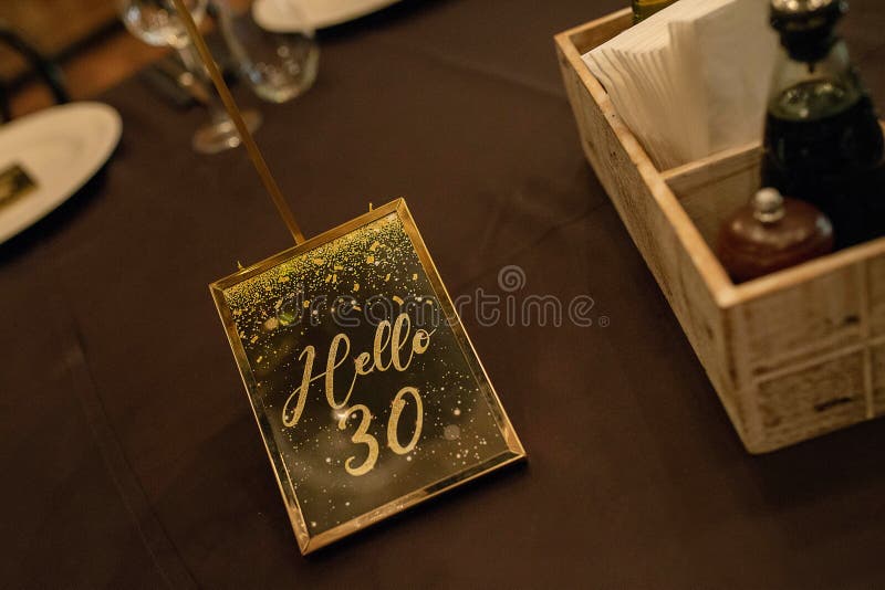 Invitation in gold and black to a 30th years birthday party framed in gold that reads `Hello 30` set on a table, elegant decoration at a restaurant prepared for the celebration of somebody turning 30.