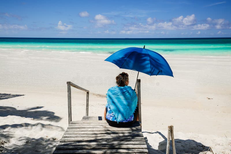 Inviterande sikt av stranden och stillsamt, turkoshav med kvinnasammanträde i förgrund, hållande paraply arkivfoto Inviterande sikt av stranden och stillsamt, turkoshav med kvinnasammanträde i förgrund, hållande paraply arkivfoto