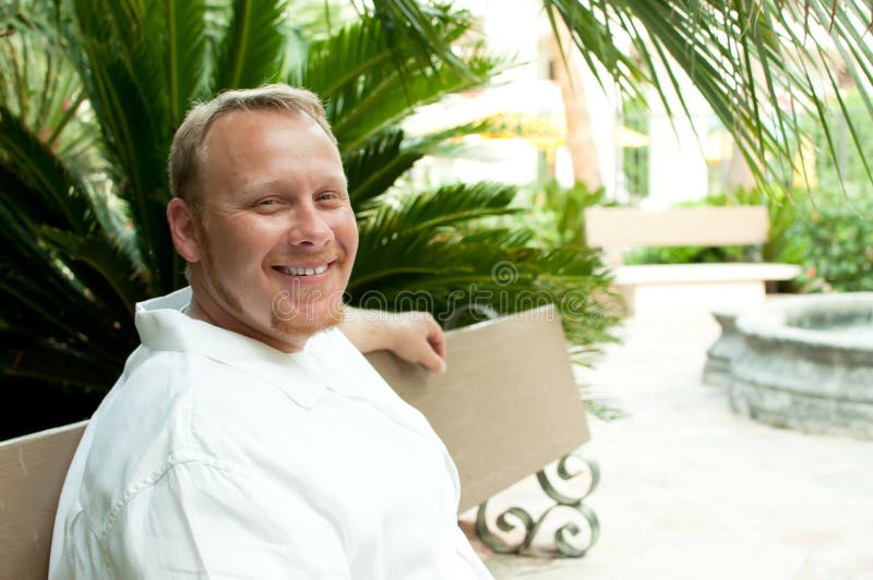 White Irish man with red hair looking at viewer with blue eyes with tropical foliage surrounding him. He is smiling and his arm is on the stone bench. White Irish man with red hair looking at viewer with blue eyes with tropical foliage surrounding him. He is smiling and his arm is on the stone bench.