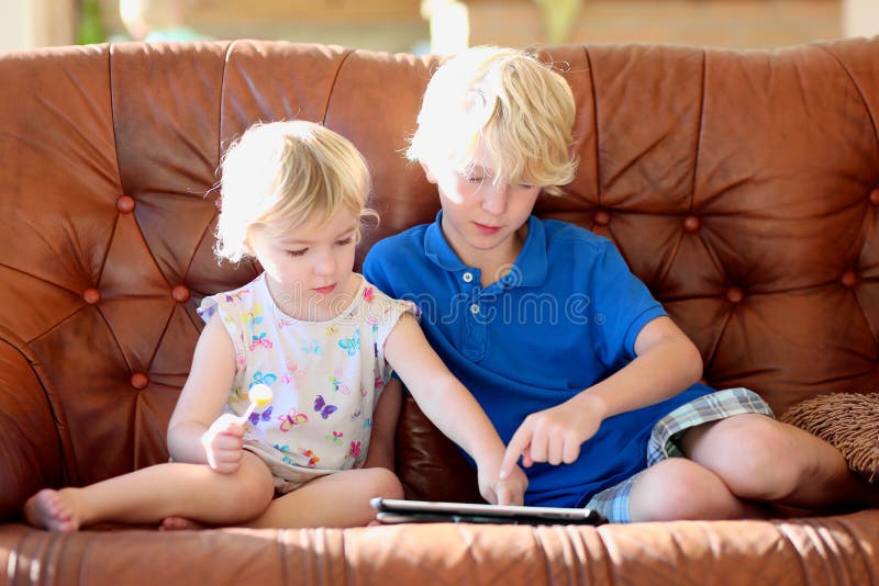 Two happy blonde kids, brother and sister, teenage boy and cute toddler girl, playing together with tablet pc sitting indoors at home in sunny living room on brown leather sofa. Two happy blonde kids, brother and sister, teenage boy and cute toddler girl, playing together with tablet pc sitting indoors at home in sunny living room on brown leather sofa