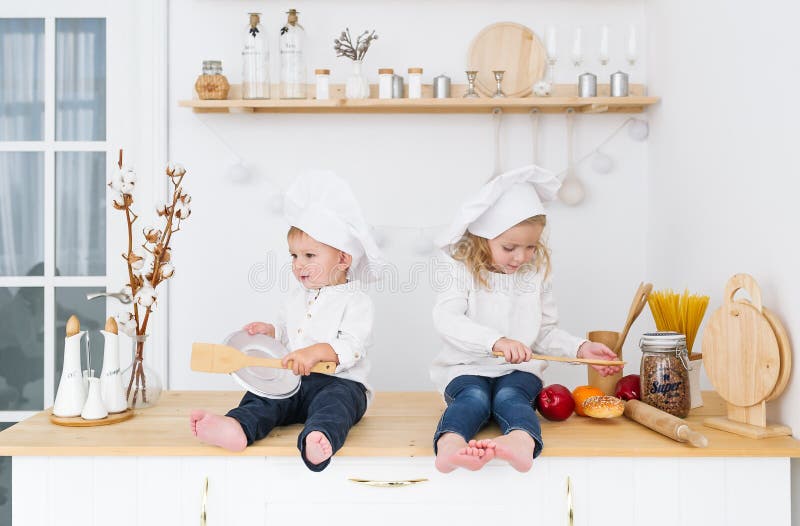 Irmão mais novo e irmã nos tampões do cozinheiro que sentam-se na cozinha na casa Os ajudantes da mãe foto de stock royalty free Irmão mais novo e irmã nos tampões do cozinheiro que sentam-se na cozinha na casa Os ajudantes da mãe foto de stock royalty free