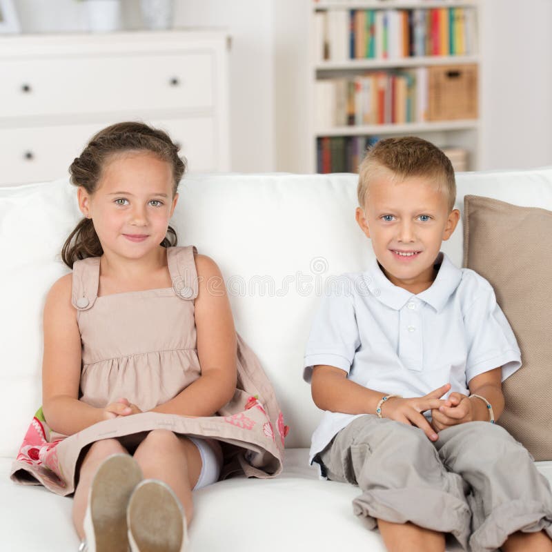 Adorable beautiful little brother and sister sitting together quietly on a couch with their hands in their laps smiling at the camera. Adorable beautiful little brother and sister sitting together quietly on a couch with their hands in their laps smiling at the camera