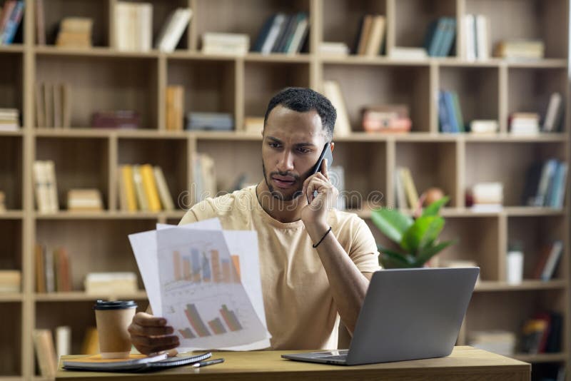 Irritated arab businessman working from home, sitting at desk with laptop, holding documents and having phone call stock photography