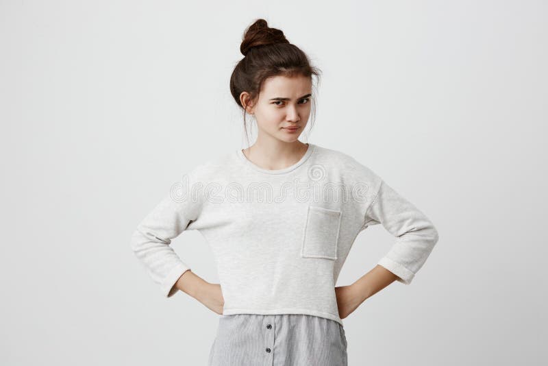 Portrait of irritated and dissatisfied brunette girl with hairbun and oval face, dark eyes, wearing loose casual sweater, frowning her eyebrows, standing with arms akimbo, being displeased with parent s words