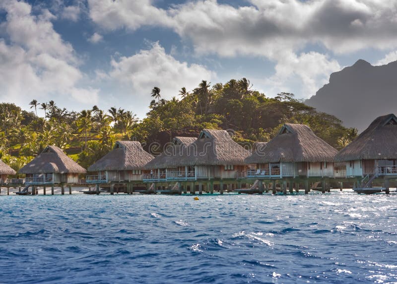 Island with palm trees and small houses on water in the ocean and mountains on a background stock photos Island with palm trees and small houses on water in the ocean and mountains on a background stock photos