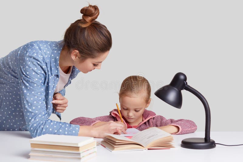 Isolated shot of young mum in fashionable shirt helps to write her small daughter, read books, do homework together, use reading l royalty free stock images Isolated shot of young mum in fashionable shirt helps to write her small daughter, read books, do homework together, use reading l royalty free stock images