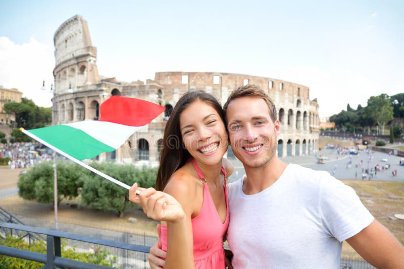 Italy travel couple with Italian flag by Colosseum embracing. Happy tourists lovers on honeymoon sightseeing having fun in front of Coliseum. Love and tourisn concept with multiracial couple.