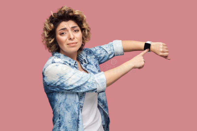 I need more time. Portrait of young pleased woman with curly hairstyle in casual blue shirt standing and pointing at her smart watch and want more time. indoor studio shot, isolated on pink background. I need more time. Portrait of young pleased woman with curly hairstyle in casual blue shirt standing and pointing at her smart watch and want more time. indoor studio shot, isolated on pink background