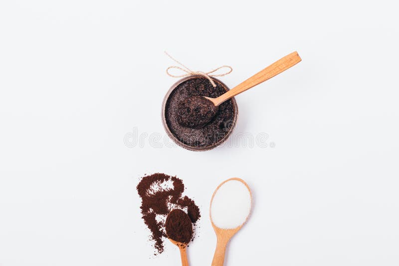 Jar of natural coffee body scrub with coconut oil next to spoons of ground beans and sugar, top view on white background