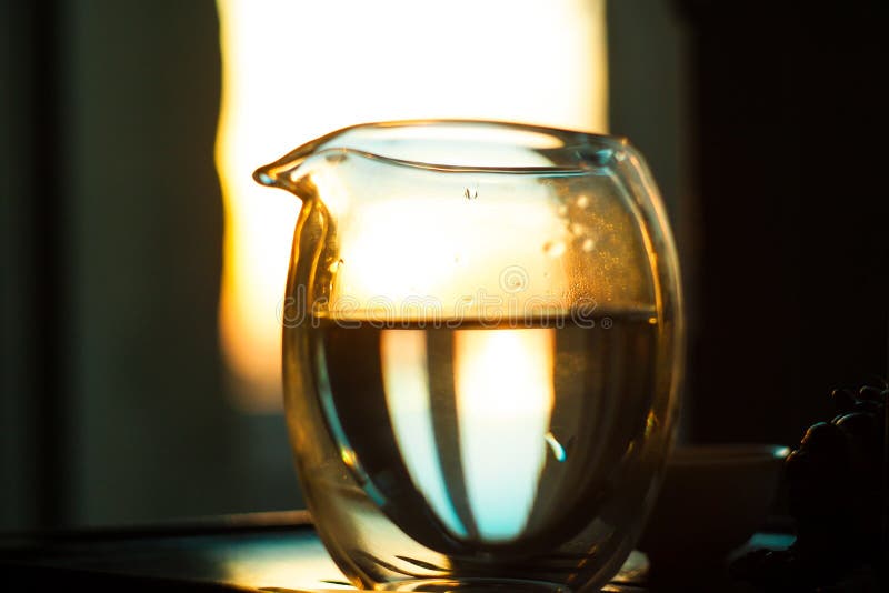 Jarra de cristal de té caliente en la tabla de madera en el fondo de la puesta del sol hermosa con los rayos del sol foto de archivo Jarra de cristal de té caliente en la tabla de madera en el fondo de la puesta del sol hermosa con los rayos del sol foto de archivo