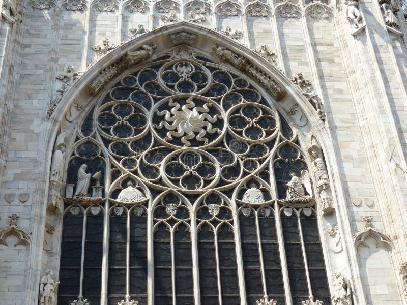 One of the decorated exterior windows of the Milan cathedral in Italy. Travel destination. Sunny day. White marble decorated walls. Neo- Gothic style. Large arched window. Some small statues  around the window and on small niches. Religious scenes and figures. Rose window. Marmoreal bas-relief. Raza, kind of sun in the center. Symbol of Sol justitia, emblem of Jesus. Heritage, culture, art and spirituality. One of the decorated exterior windows of the Milan cathedral in Italy. Travel destination. Sunny day. White marble decorated walls. Neo- Gothic style. Large arched window. Some small statues  around the window and on small niches. Religious scenes and figures. Rose window. Marmoreal bas-relief. Raza, kind of sun in the center. Symbol of Sol justitia, emblem of Jesus. Heritage, culture, art and spirituality.