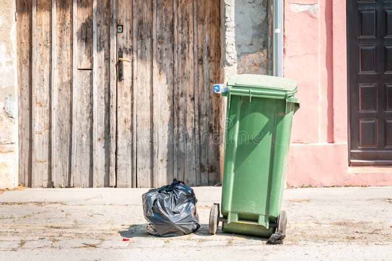 One single green garbage can and black plastic junk bag on the street in the city waiting for dumper truck to collect in front of the house. One single green garbage can and black plastic junk bag on the street in the city waiting for dumper truck to collect in front of the house.