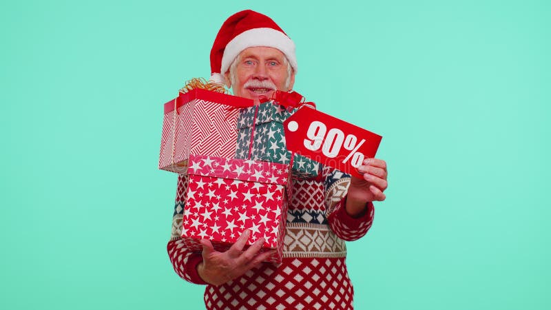 Senior grandfather man in Christmas sweater showing gift box and 90 Percent discount inscriptions banner text. Black Friday. Good holiday low prices for shopping. Blue studio wall background. New Year. Senior grandfather man in Christmas sweater showing gift box and 90 Percent discount inscriptions banner text. Black Friday. Good holiday low prices for shopping. Blue studio wall background. New Year