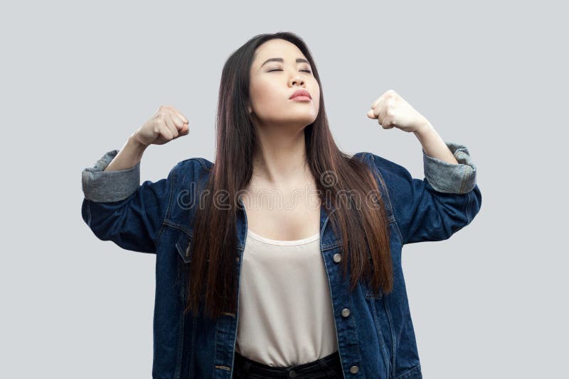 I am strong and independent. Portrait of proud satisfied beautiful brunette asian young woman in casual blue jacket and makeup standing with raised arms. studio shot, isolated on light grey background. I am strong and independent. Portrait of proud satisfied beautiful brunette asian young woman in casual blue jacket and makeup standing with raised arms. studio shot, isolated on light grey background