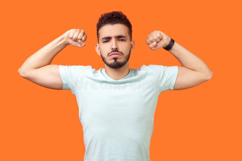 I am strong and powerful. Portrait of confident successful brunette man with beard in white t-shirt showing biceps, full of motivation and willpower. indoor studio shot  on orange background. I am strong and powerful. Portrait of confident successful brunette man with beard in white t-shirt showing biceps, full of motivation and willpower. indoor studio shot  on orange background