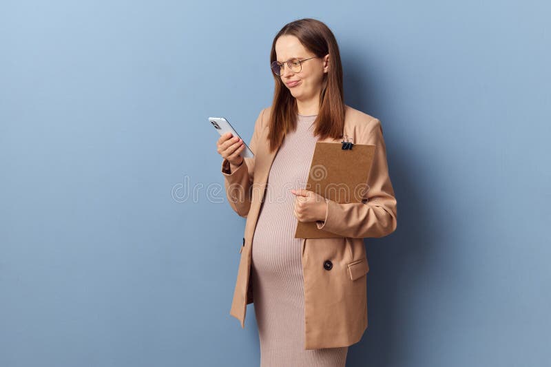 Serious young adult pregnant woman wearing dress and jacket posing with clipboard in hands using smartphone for business inline communication via chat isolated over blue background. Serious young adult pregnant woman wearing dress and jacket posing with clipboard in hands using smartphone for business inline communication via chat isolated over blue background