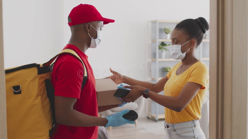Jeune afro-américaine en masque facial protecteur payant avec smartphone et obtenir le paquet de commande du courrier banque de vidéos