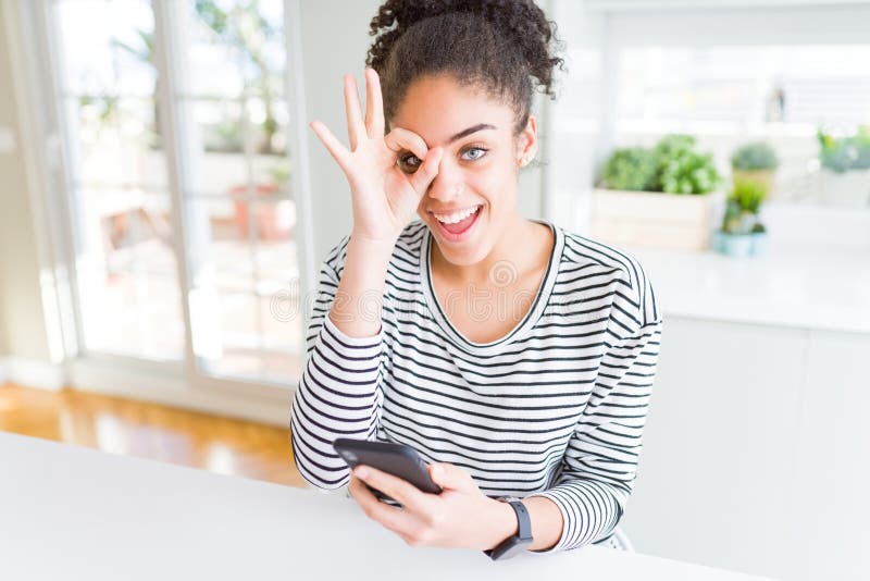 Jeune afro-américaine utilisant smartphone textos un message avec un visage heureux sourire faire ok signe avec main sur l'oeil re photo libre de droits