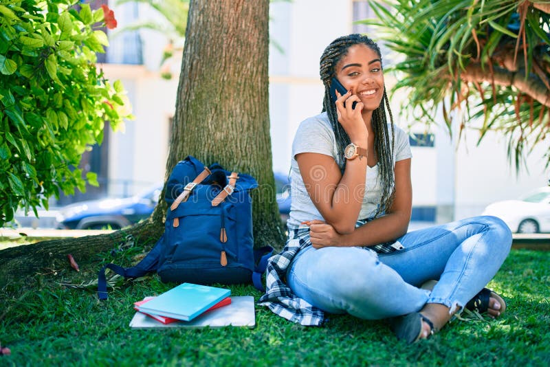 Jeune afro-américaine étudiante souriante joyeuse parlant sur smartphone assise sur l'herbe du campus universitaire photo stock