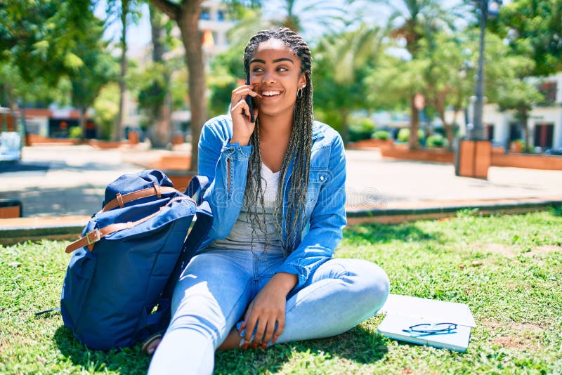 Jeune afro-américaine étudiante souriante joyeuse parlant sur smartphone assise sur l'herbe du campus universitaire image stock