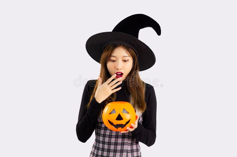 Young asian pretty woman in black halloween costume wearing witch hat and holding pumpkin lantern posing surprised and excited on white background. Young asian pretty woman in black halloween costume wearing witch hat and holding pumpkin lantern posing surprised and excited on white background.