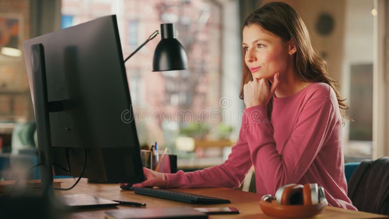 Young Beautiful Adult Woman in Pink Jumper Working from Home on Desktop Computer. Creative Female Checking and Writing Emails. Loft Apartment with Urban City View from Big Window. Young Beautiful Adult Woman in Pink Jumper Working from Home on Desktop Computer. Creative Female Checking and Writing Emails. Loft Apartment with Urban City View from Big Window.