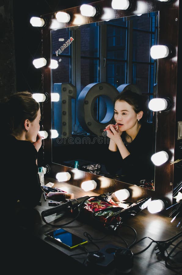 Jeune belle femme appliquant ses lèvres de maquillage, regardant dans un miroir, se reposant sur la chaise le vestiaire de théâtr photos libres de droits Jeune belle femme appliquant ses lèvres de maquillage, regardant dans un miroir, se reposant sur la chaise le vestiaire de théâtr photos libres de droits