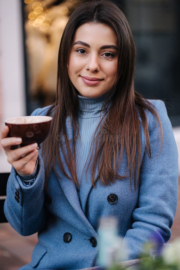 Jeune belle femme assise à l'extérieur dans le café et boire du café. femme heureuse en manteau bleu d'hiver. humeur de noël photos stock