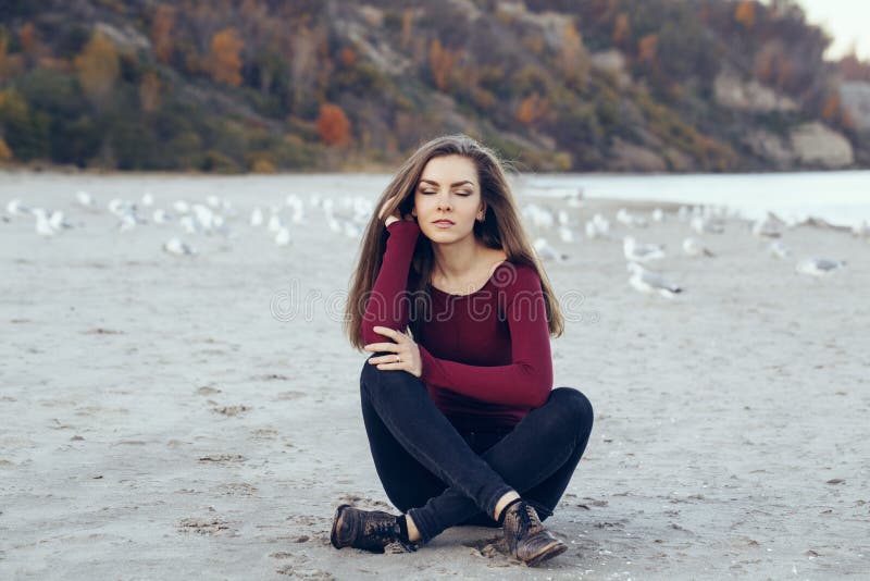 Jeune belle femme avec les yeux fermés, longs cheveux, jeans noirs de port et chemise rouge, se reposant sur le sable sur la plag photos libres de droits Jeune belle femme avec les yeux fermés, longs cheveux, jeans noirs de port et chemise rouge, se reposant sur le sable sur la plag photos libres de droits