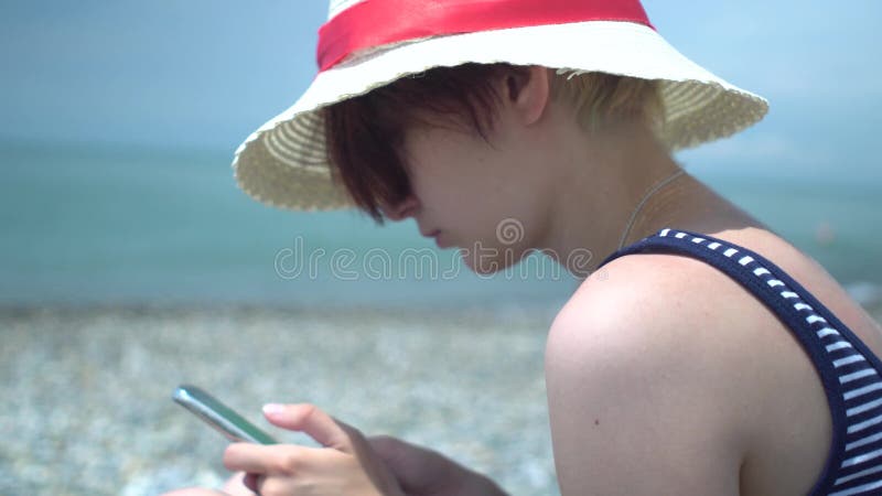 Jeune belle femme de hippie à l'aide du smartphone utilisant le rétro chapeau et le maillot de bain avec les rayures bleues et bl