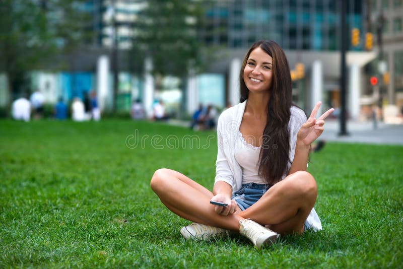 Jeune belle femme de touristes caucasienne s'asseyant sur l'herbe verte au parc de ville et montrant le signe de la victoire v