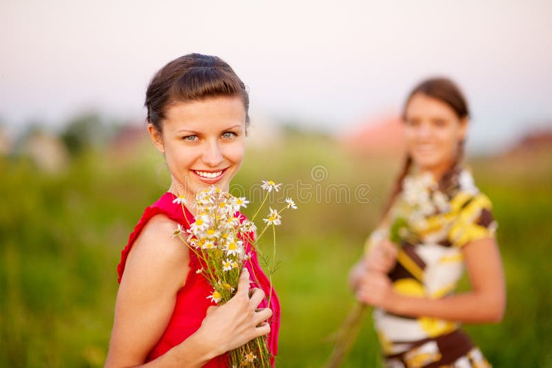 Two young beautiful woman wllith bouquet of camomiles against a summer landscape. Two young beautiful woman wllith bouquet of camomiles against a summer landscape.