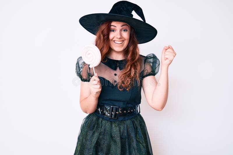 Young beautiful woman wearing witch halloween costume holding lollipop screaming proud, celebrating victory and success very excited with raised arms. Young beautiful woman wearing witch halloween costume holding lollipop screaming proud, celebrating victory and success very excited with raised arms