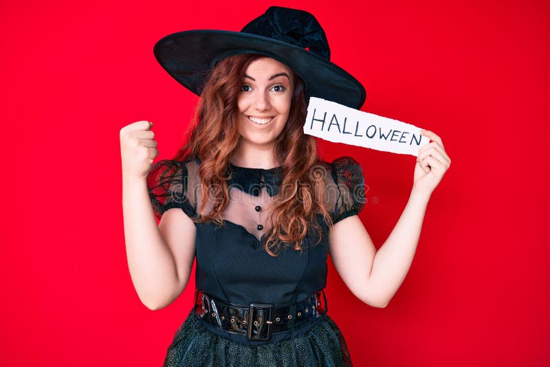 Young beautiful woman wearing witch costume holding paper with halloween message screaming proud, celebrating victory and success very excited with raised arms. Young beautiful woman wearing witch costume holding paper with halloween message screaming proud, celebrating victory and success very excited with raised arms