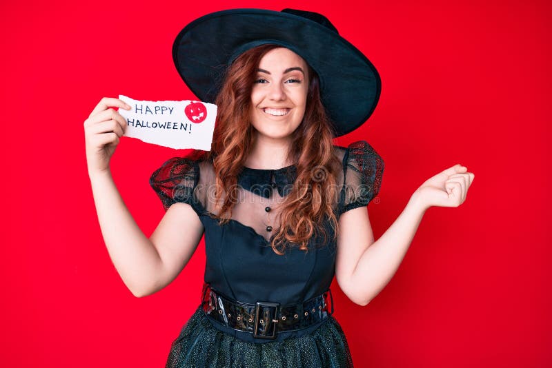 Young beautiful woman wearing witch costume holding happy halloween message paper screaming proud, celebrating victory and success very excited with raised arm. Young beautiful woman wearing witch costume holding happy halloween message paper screaming proud, celebrating victory and success very excited with raised arm