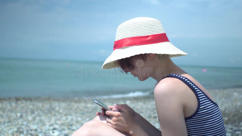 Jeune belle femme utilisant le rétro chapeau et le maillot de bain avec les rayures bleues et blanches en plage isolée d'été util