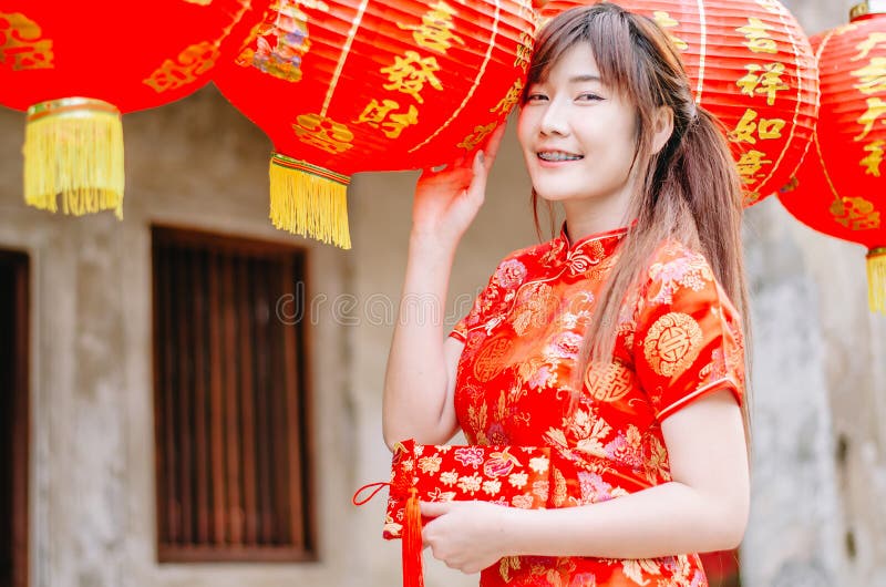 Jeune belle fille asiatique dans le support rouge traditionnel chinois de sourire de robe près de la lampe de porcelaine, par nou photo stock Jeune belle fille asiatique dans le support rouge traditionnel chinois de sourire de robe près de la lampe de porcelaine, par nou photo stock