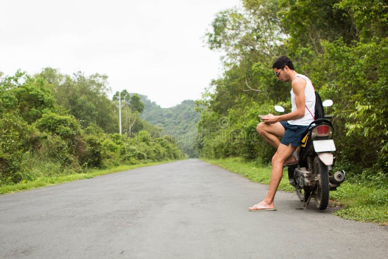 Jeune cavalier de touristes s'asseyant sur une motocyclette de location utilisant un smartphone au milieu d'une route image libre de droits Jeune cavalier de touristes s'asseyant sur une motocyclette de location utilisant un smartphone au milieu d'une route image libre de droits