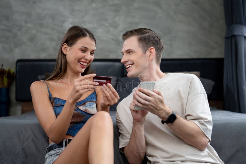 Young couple sit in the home bedroom using online payment app and digital wallet on smartphone to pay with credit card. E commerce shopping and modern purchasing via mobile internet. Unveiling. Young couple sit in the home bedroom using online payment app and digital wallet on smartphone to pay with credit card. E commerce shopping and modern purchasing via mobile internet. Unveiling