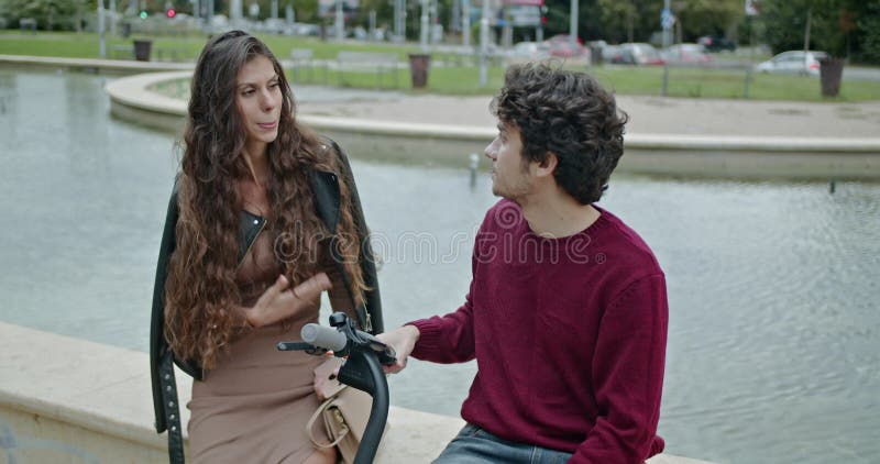 Jeune couple parlant et riant près d'une fontaine dans un parc avec un scooter électrique Profitez d'une journée ensoleillée en pl banque de vidéos