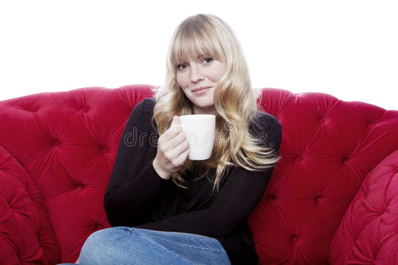 Jeune cuvette de boissons de fille de café d'une chevelure blonde sur le sofa rouge dans l'avant photographie stock libre de droits Jeune cuvette de boissons de fille de café d'une chevelure blonde sur le sofa rouge dans l'avant photographie stock libre de droits