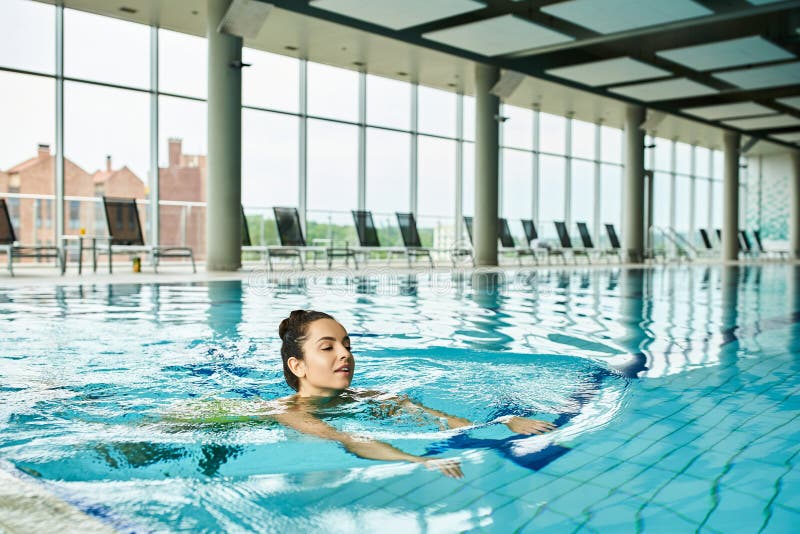 A young and beautiful brunette woman peacefully swims in a large indoor pool at a luxurious spa, stock photo. A young and beautiful brunette woman peacefully swims in a large indoor pool at a luxurious spa, stock photo