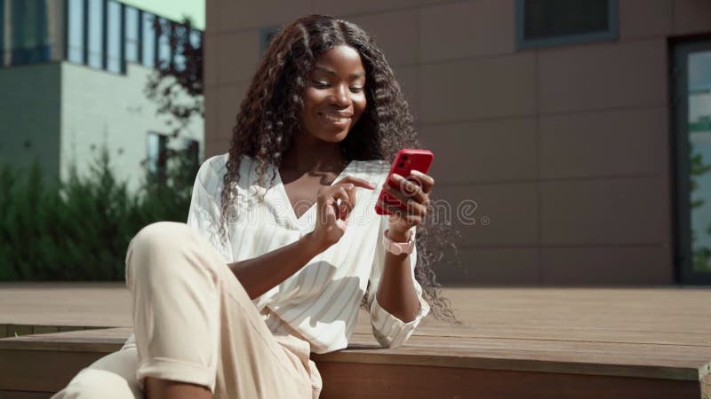 Jeune femme africaine noire heureuse tenant un smartphone utilisant des applications mobiles extérieur. banque de vidéos
