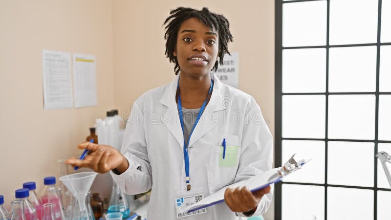 Jeune femme afro-américaine avec des cadenas habillés en technicien de laboratoire tenant un presse-papiers dans un cadre de labor image stock