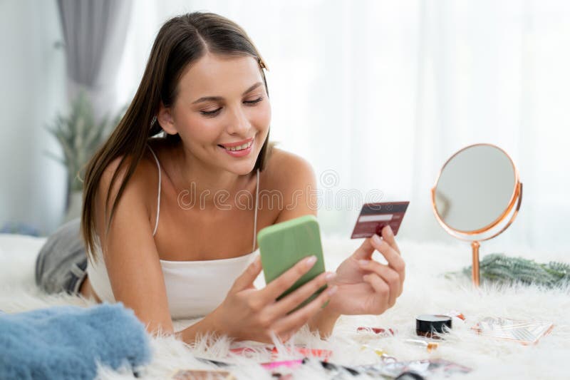 Young woman lying on the bed using online payment app and digital wallet on smartphone to pay with credit card. E commerce shopping and modern purchasing via mobile internet. Unveiling. Young woman lying on the bed using online payment app and digital wallet on smartphone to pay with credit card. E commerce shopping and modern purchasing via mobile internet. Unveiling
