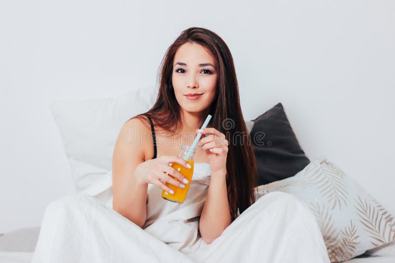 Jeune femme asiatique de sourire heureuse de fille de beaux longs cheveux s'asseyant dans le lit avec le jus d'orange dans le pot photo stock Jeune femme asiatique de sourire heureuse de fille de beaux longs cheveux s'asseyant dans le lit avec le jus d'orange dans le pot photo stock
