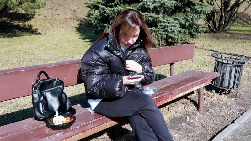 Jeune femme assise sur le banc du parc en vérifiant le téléphone tout en dégustant une collation par temps ensoleillé banque de vidéos