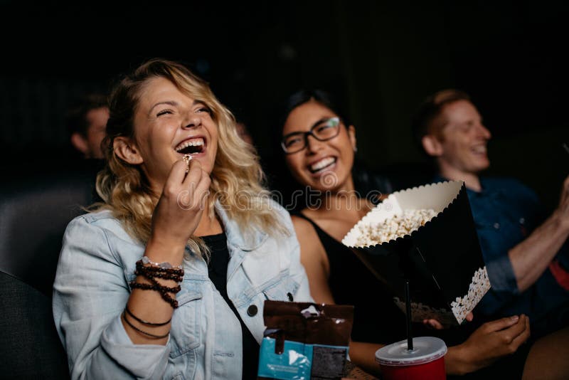 Young women with friends watching movie in cinema and laughing. Group of people in theater with popcorns and drinks. Young women with friends watching movie in cinema and laughing. Group of people in theater with popcorns and drinks.