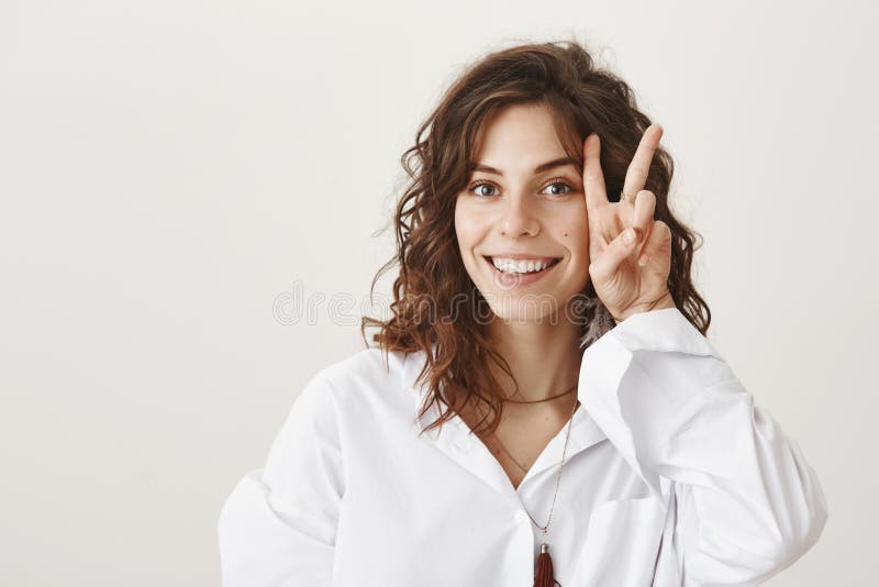 Jeune femme avec du charme avec les cheveux bouclés mignons montrant le geste de paix ou de victoire près du visage tout en souri photos stock Jeune femme avec du charme avec les cheveux bouclés mignons montrant le geste de paix ou de victoire près du visage tout en souri photos stock