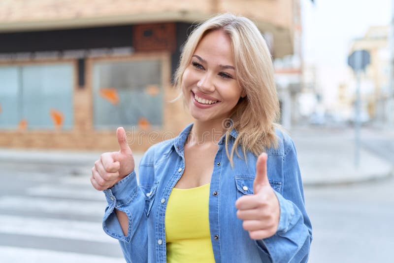 Jeune femme blonde souriante confiante faire ok signe avec des pouces dans la rue image libre de droits